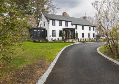 A large, white colonial house with white siding, black windows, and a light wooden door, surrounded by green grass and various flowers.