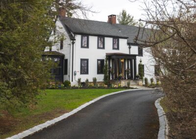 A large, white colonial house with white siding, black windows, and a light wooden door, surrounded by green grass and various flowers.