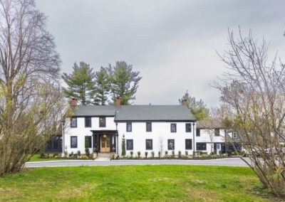 A large, white colonial house with white siding, black windows, and a light wooden door, surrounded by green grass and various flowers.