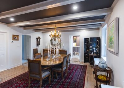 A spacious dining room featuring a large table with six chairs around it, an elegant chandelier above, and a deep red and blue intricate carpet underneath.