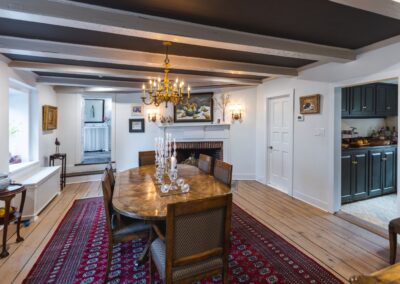 A spacious dining room featuring a large table with six chairs around it, an elegant chandelier above, a deep red and blue intricate carpet, and a fireplace on the other end.