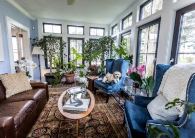 The colorful sun room, featuring a plethora of windows, large plants, two deep blue chairs opposite a small brown leather couch, with a small glass table in the middle. There is a cute dog sitting on the farther chair.
