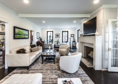A large living room with white walls and floors, featuring white couches and chairs, with a textured black & white carpet, plenty of art on the walls, and a fireplace with a tv sitting above it.