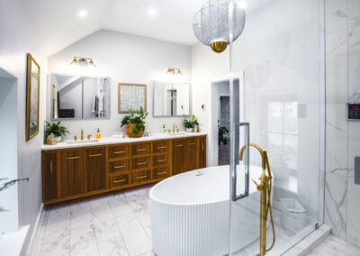 A bright, white primary bathroom with a large stand-alone tub, two sinks, stunning chandelier, and wooden textured cabinets.
